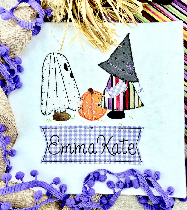 May include: A white fabric panel with an embroidered design of a ghost, a witch, and a pumpkin. The witch is wearing a black hat and a colorful striped dress. The panel has a blue and white gingham banner with the name "Emma Kate" embroidered on it.