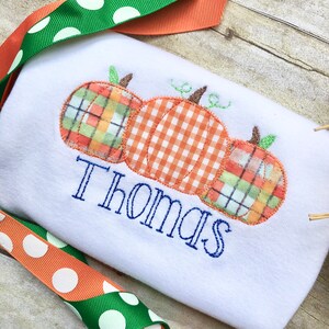 May include: White onesie with three embroidered pumpkins in orange, green, and plaid. The name "Thomas" is embroidered below the pumpkins.
