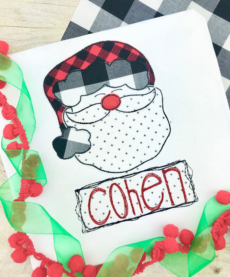 May include: White fabric with an embroidered Santa Claus face. The Santa face features a red and black buffalo plaid hat, a red nose, and a dotted beard. The name "COHEN" is embroidered in red within a black outlined rectangle.