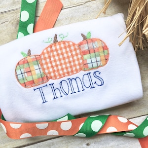 May include: White onesie with three embroidered pumpkins in orange, green, and plaid. The name "Thomas" is embroidered below the pumpkins.