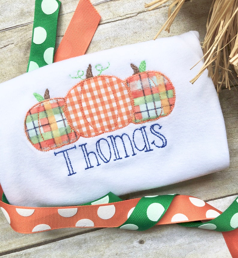 May include: White onesie with three embroidered pumpkins in orange, green, and plaid. The name "Thomas" is embroidered below the pumpkins.