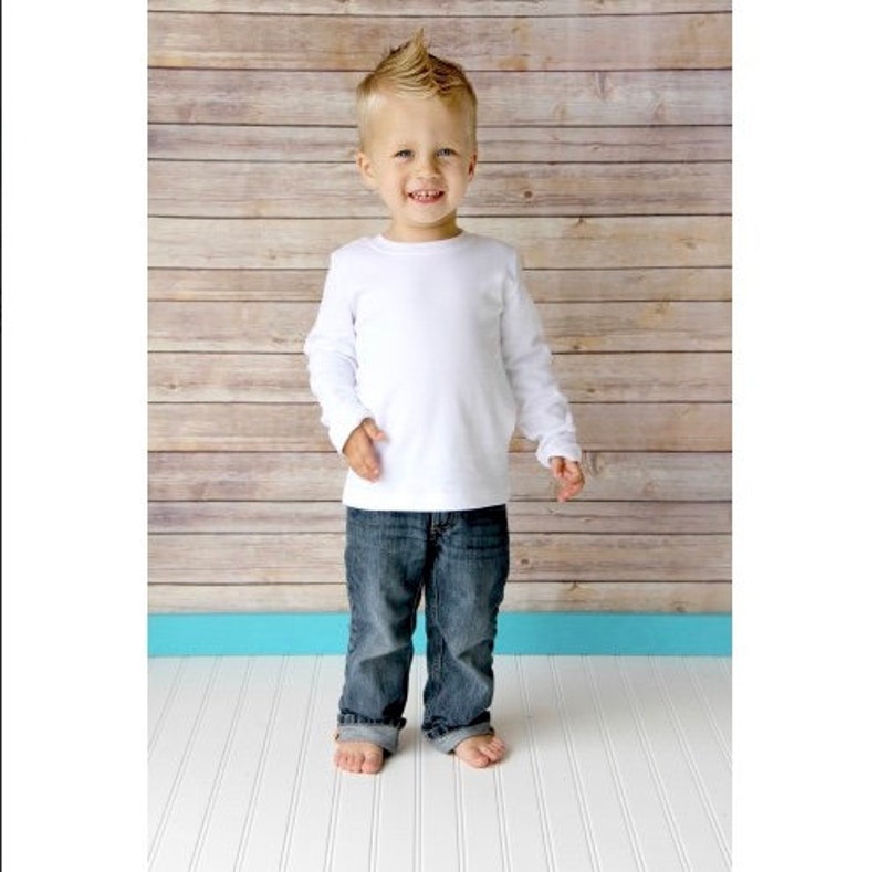 May include: A young boy wearing a white long-sleeved shirt and blue jeans stands in front of a wood panel background.