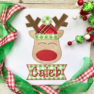 May include: A white onesie with a red and green plaid reindeer applique. The reindeer has a green and white pom-pom hat and a red nose. The onesie has the name "Caleb" embroidered on a green and white plaid banner.