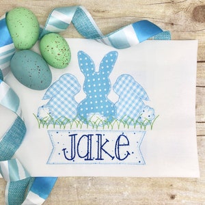 May include: A white embroidered appliqu&eacute; of three blue bunnies with a banner that reads "Jake". The bunnies are in a row, with the middle bunny being the largest. The bunnies are all wearing different patterns, including polka dots and gingham. The banner is decorated with small dots.