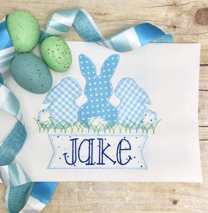 May include: A white embroidered appliqu&eacute; of three blue bunnies with a banner that reads "Jake". The bunnies are in a row, with the middle bunny being the largest. The bunnies are all wearing different patterns, including polka dots and gingham. The banner is decorated with small dots.