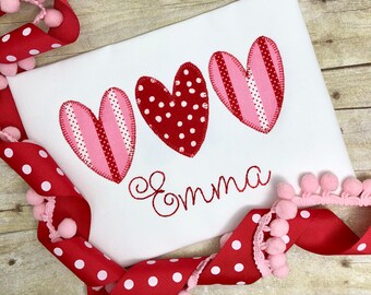 May include: White t-shirt with three red and pink hearts and the name "Emma" embroidered in red.