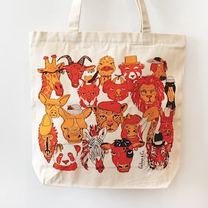 May include: A beige canvas tote bag with a colorful illustration of various animal faces. The animals are drawn in a cartoon style and include a giraffe, a goat, a rhino, a zebra, a panda, a tiger, and a lion. The bag has a short handle and is perfect for carrying everyday essentials.