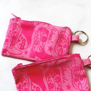 May include: Two pink zippered pouches with a cowboy boot pattern and the text "Calhoun & Co." printed on them. Each pouch has a silver key ring attached.