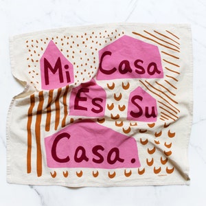 May include: A white tea towel with a pink and brown geometric design. The text "Mi Casa Es Su Casa." is printed on the towel.