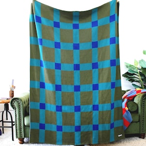 May include: A rectangular knit throw blanket featuring a grid pattern of olive green squares separated by intersecting bands of teal and bright blue. The blanket has a small white rectangular tag attached to the bottom right corner.