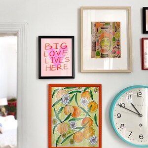 May include: A white wall with a collection of framed artwork and a clock. The artwork includes a print with the text "Big Love Lives Here" and a print of a woman with a variety of hair care products. The clock is blue with white numbers and hands.