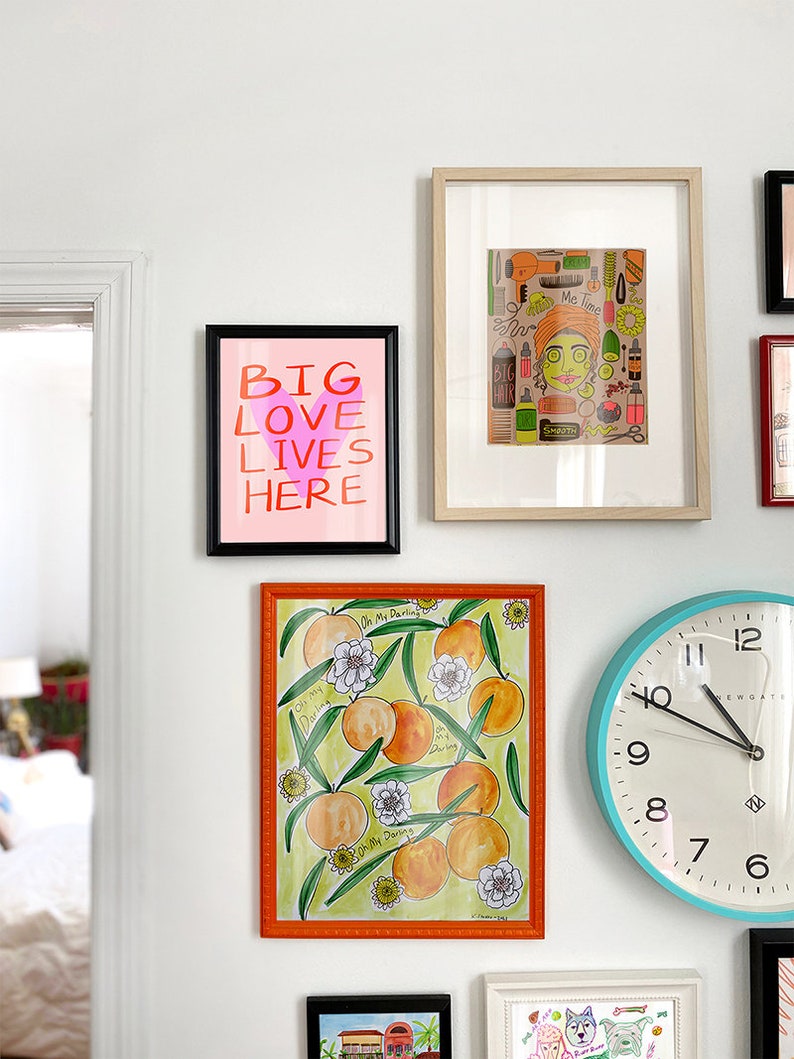 May include: A white wall with a collection of framed artwork and a clock. The artwork includes a print with the text "Big Love Lives Here" and a print of a woman with a variety of hair care products. The clock is blue with white numbers and hands.