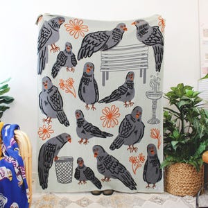 May include: A light green throw blanket featuring a repeating pattern of gray pigeons, orange flowers, a park bench, and a fountain. The blanket is displayed in a well-lit room with other decorative items.
