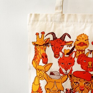 May include: A white canvas tote bag with a colorful illustration of various animals wearing hats and accessories. The animals include a giraffe, a goat, a fox, a rhino, a zebra, a panda, and a monkey.