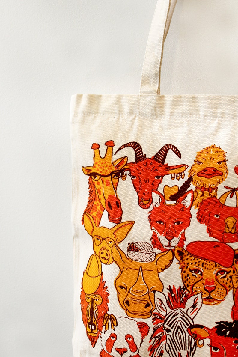 May include: A white canvas tote bag with a colorful illustration of various animals wearing hats and accessories. The animals include a giraffe, a goat, a fox, a rhino, a zebra, a panda, and a monkey.