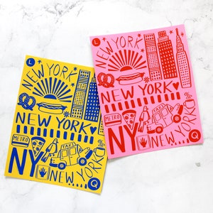 May include: Two illustrated prints of New York City landmarks, one in yellow with blue details and the other in pink with red details. Both prints feature the text "NEW YORK" and include iconic symbols like hot dogs, pretzels, and taxis.