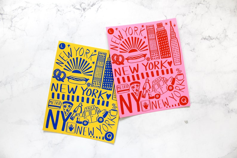 May include: Two illustrated prints of New York City landmarks, one in yellow with blue details and the other in pink with red details. Both prints feature the text "NEW YORK" and include iconic symbols like hot dogs, pretzels, and taxis.