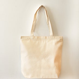 May include: A plain, off-white canvas tote bag with two long handles. The bag is hanging on a white wall.