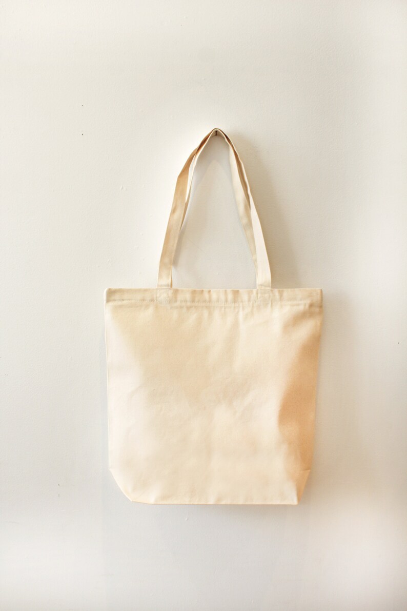 May include: A plain, off-white canvas tote bag with two long handles. The bag is hanging on a white wall.
