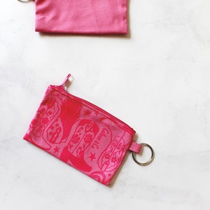 May include: Two pink zippered pouches with key rings. The top pouch is plain and the bottom pouch has a pink floral design.
