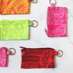 May include: Nine colourful zippered pouches with different patterns and designs. The pouches have a key ring attached. The patterns include a martini glass, cowboy boots, a seashell, a food market, and a stick of butter.