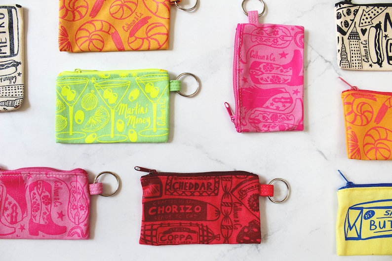 May include: Nine colourful zippered pouches with different patterns and designs. The pouches have a key ring attached. The patterns include a martini glass, cowboy boots, a seashell, a food market, and a stick of butter.