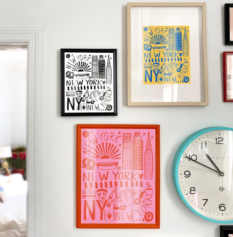 May include: Three framed prints with a New York City theme. The prints are in black and white, blue and yellow, and pink and orange. Each print features a different design with iconic New York City symbols like the skyline, hot dogs, pretzels, and taxis.