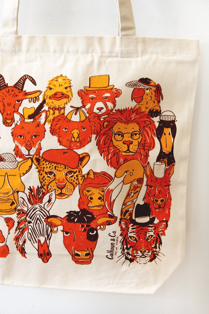 May include: A white canvas tote bag with a colorful illustration of various animals wearing hats. The animals include a lion, a tiger, a zebra, a cow, a monkey, a duck, a koala, a goat, a fox, a llama, a cheetah, a rhinoceros, and a parrot. The text "Calhoun & Co. NEW YORK" is printed on the bag.