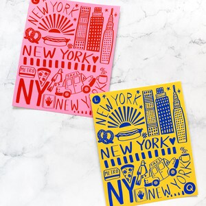 May include: Two illustrated prints of New York City landmarks, one in red on a pink background and one in blue on a yellow background. Both prints feature the text "New York" and include iconic symbols like the Empire State Building, hot dogs, pretzels, and taxis.