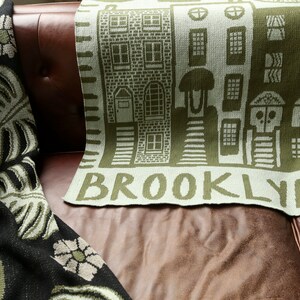May include: A green and white knitted throw blanket with a design of brown buildings and the word "BROOKLYN" in green letters.