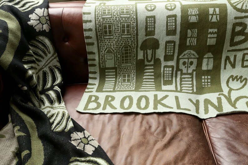 May include: A green and white knitted throw blanket with a design of brown buildings and the word "BROOKLYN" in green letters.