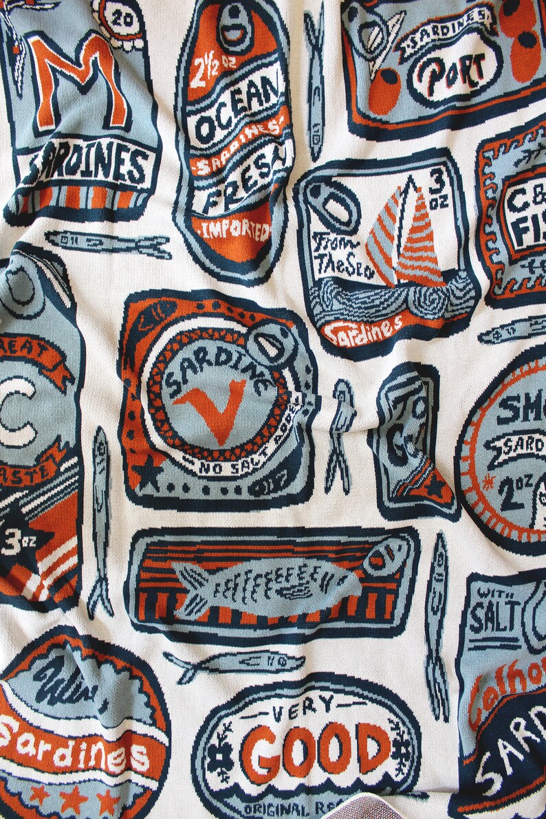 May include: A colorful patterned fabric with a repeating design of sardine cans. The cans are in various colors, including blue, orange, and red, and feature different text, such as "Ocean Fresh Sardines" and "Very Good".