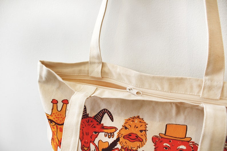 May include: A beige canvas tote bag with a zipper closure. The bag features a colorful print of various animals, including a giraffe, a goat, an ostrich, and a bear.