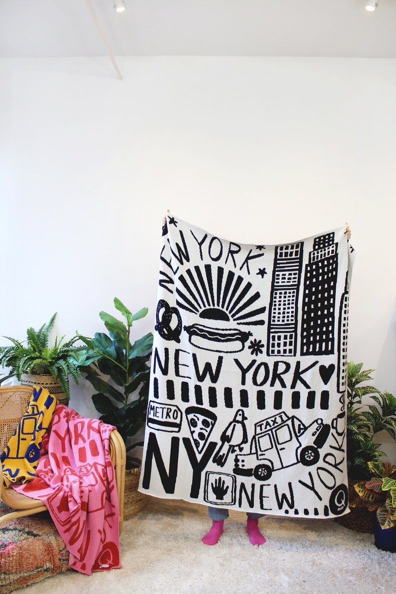 May include: A white throw blanket with black illustrations of New York City landmarks, food, and transportation. The blanket features the words "New York" and "NY" in a bold, black font.