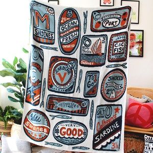 May include: A white throw blanket with a colorful pattern of sardine cans. The cans have various labels with text such as "Ocean Sardines", "Wild Sardines", "Very Good", and "Calhoun's Sardine".