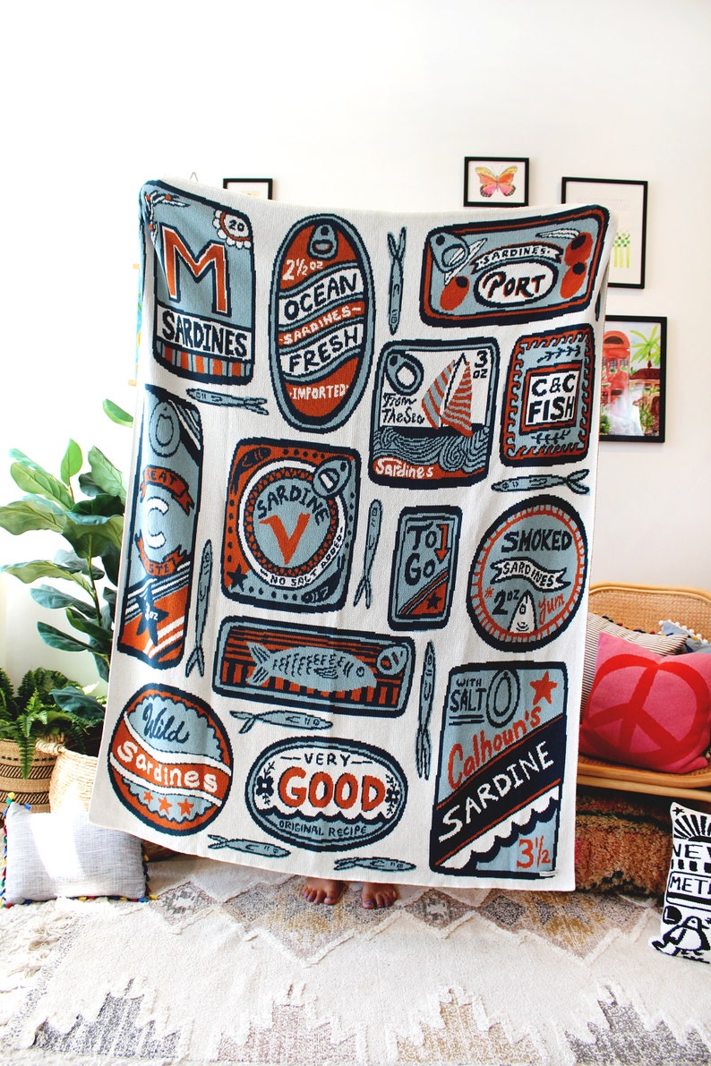 May include: A white throw blanket with a colorful pattern of sardine cans. The cans have various labels with text such as "Ocean Sardines", "Wild Sardines", "Very Good", and "Calhoun's Sardine".