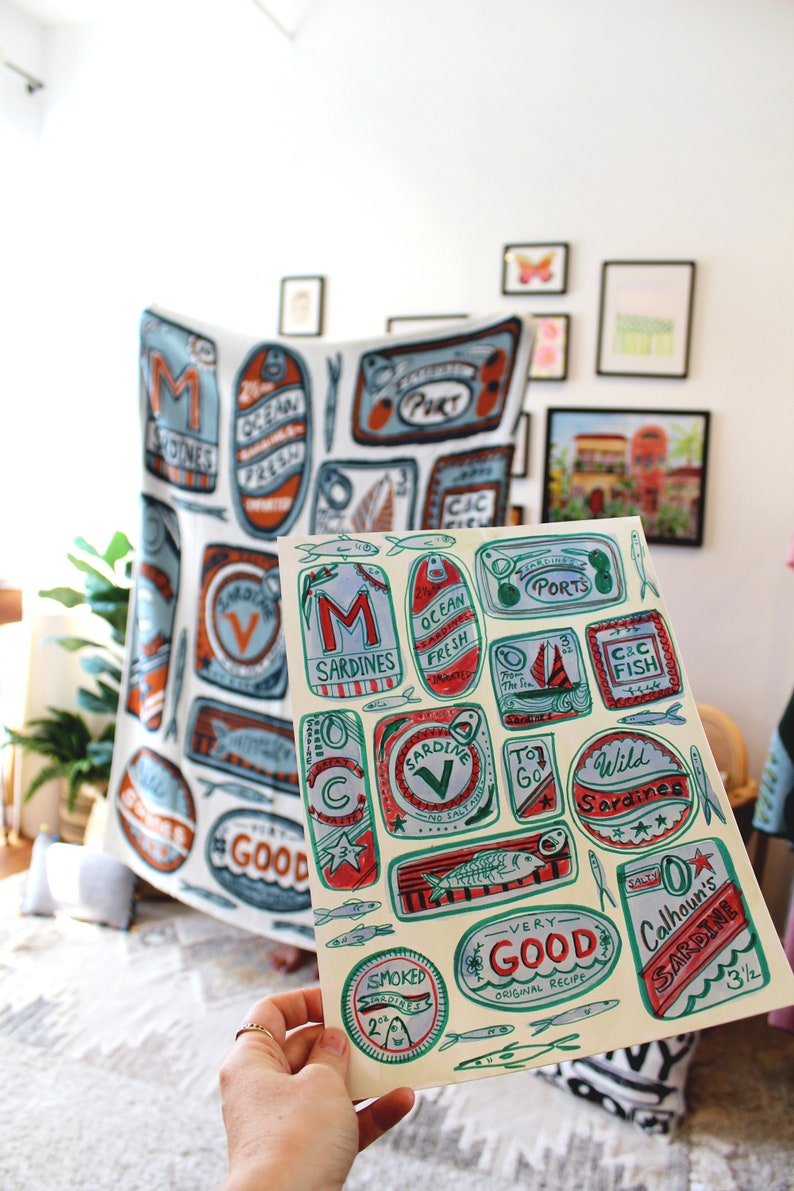 May include: A sheet of paper with colorful illustrations of sardine cans. The cans have different designs and text, including "Ocean Fresh", "To Go", "Wild Sardines", "Very Good", and "Calhoun's Sardine".