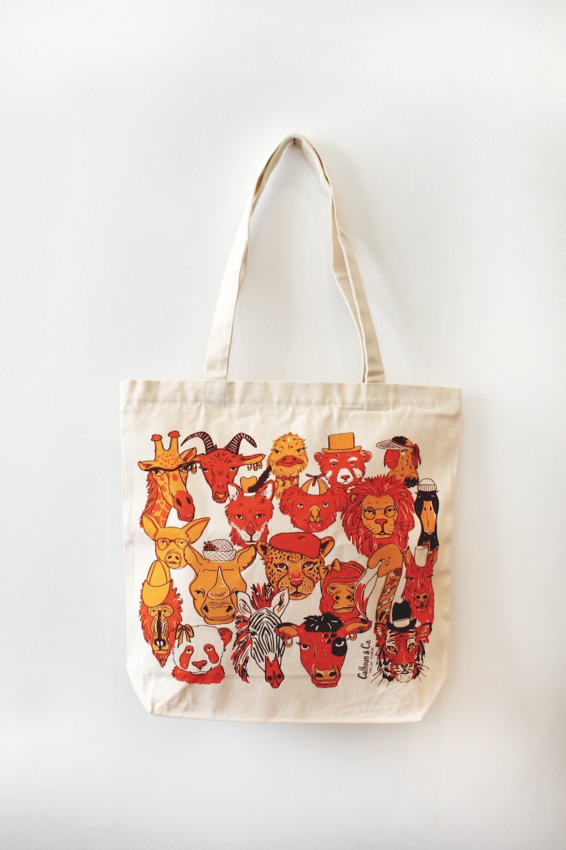 May include: A beige canvas tote bag with a colorful illustration of various animal faces. The animals are drawn in a cartoon style and include a giraffe, a goat, a rhino, a zebra, a panda, a tiger, and a lion. The bag has a short handle and is perfect for carrying everyday essentials.