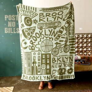 May include: A green and white woven throw blanket with a graphic design featuring the word "Brooklyn" and various illustrations of city life, including buildings, a pizza slice, a basketball, a bicycle, and a bird.