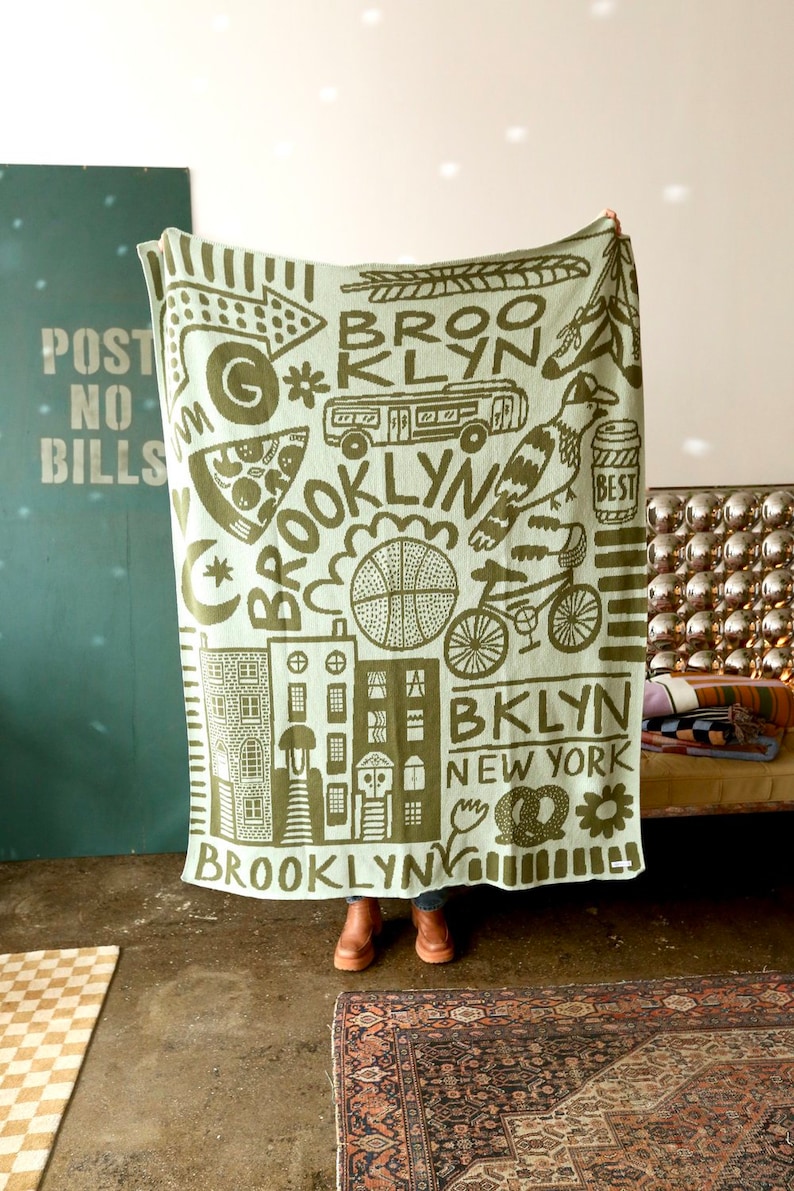 May include: A green and white woven throw blanket with a graphic design featuring the word "Brooklyn" and various illustrations of city life, including buildings, a pizza slice, a basketball, a bicycle, and a bird.