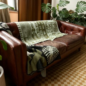 May include: A brown leather sofa with a green and black throw blanket featuring the text "Brooklyn, New York". The sofa is in a living room with a potted plant and a checkered rug.