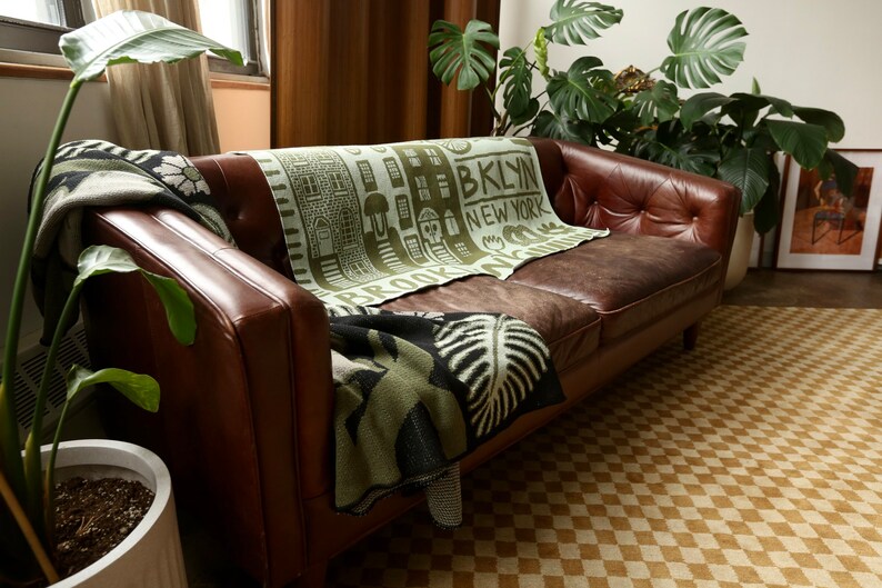 May include: A brown leather sofa with a green and black throw blanket featuring the text "Brooklyn, New York". The sofa is in a living room with a potted plant and a checkered rug.