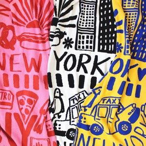 May include: Three colorful knitted blankets with New York City themed designs. The blankets are pink, white, and yellow with black, blue, and red accents. The blankets feature images of a sunrise, buildings, a taxi, and the words "New York".