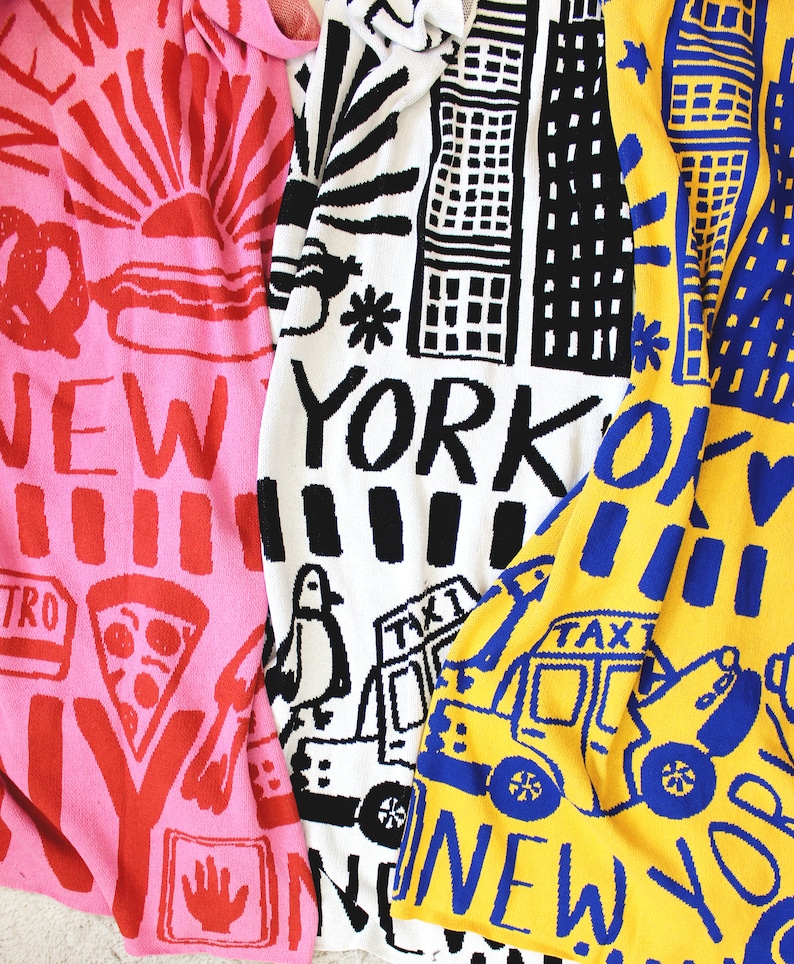May include: Three colorful knitted blankets with New York City themed designs. The blankets are pink, white, and yellow with black, blue, and red accents. The blankets feature images of a sunrise, buildings, a taxi, and the words "New York".