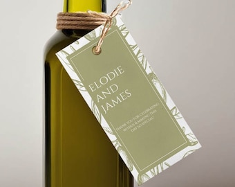 May include: A personalized olive oil bottle with a tag that says "Elodie and James". The bottle is tied with twine and has a cork stopper. The tag is decorated with a green olive branch design. The bottle is sitting on a white surface.