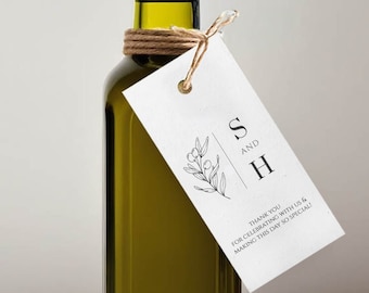 May include: A personalized olive oil bottle with a tag that says "S and H Thank you for celebrating with us & making this day so special". The bottle is tied with twine and has a cork stopper. The bottle is sitting on a white surface with a green olive branch in the foreground.