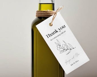 May include: A clear glass bottle of olive oil with a cork stopper and a brown twine bow. A white tag with black text reads "Thank you for celebrating with us" and "[Name] and [Name] July 10, 2023". The tag is tied to the bottle with twine. A round sticker with a white background and orange text reads "CAKE WALK COLLECTIVE".