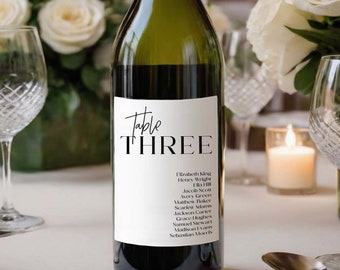 May include: A white wine bottle with a black and white label that says "Table Three" and lists a number of names. The bottle is sitting on a white tablecloth with a white rose bouquet in the background.