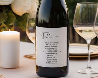 May include: A white wine bottle with a black label that says "Table Two" and lists a number of names. The bottle is sitting on a table with two wine glasses and a white candle.