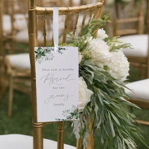 Peut inclure: Carte blanche avec une bordure de feuillage vert et le texte "This row is kindly Reserved for the groom's family." accrochée à une chaise dorée avec des fleurs blanches et de la verdure.